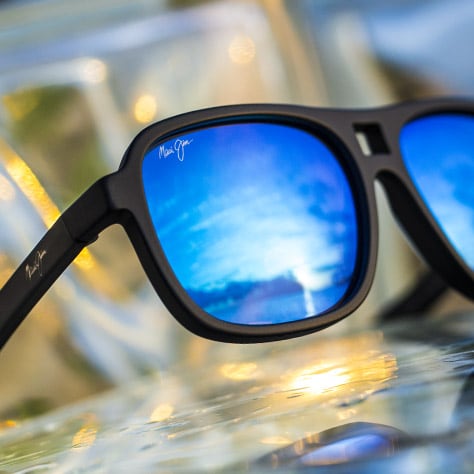 Sonnenbrille mit mattschwarzer Fassung und blauen Gläsern, in denen sich der Himmel spiegelt